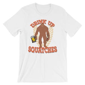 May include: White t-shirt with a brown Bigfoot holding a beer and a bottle. The text "Drink Up Squatchies" is printed in orange on the shirt.