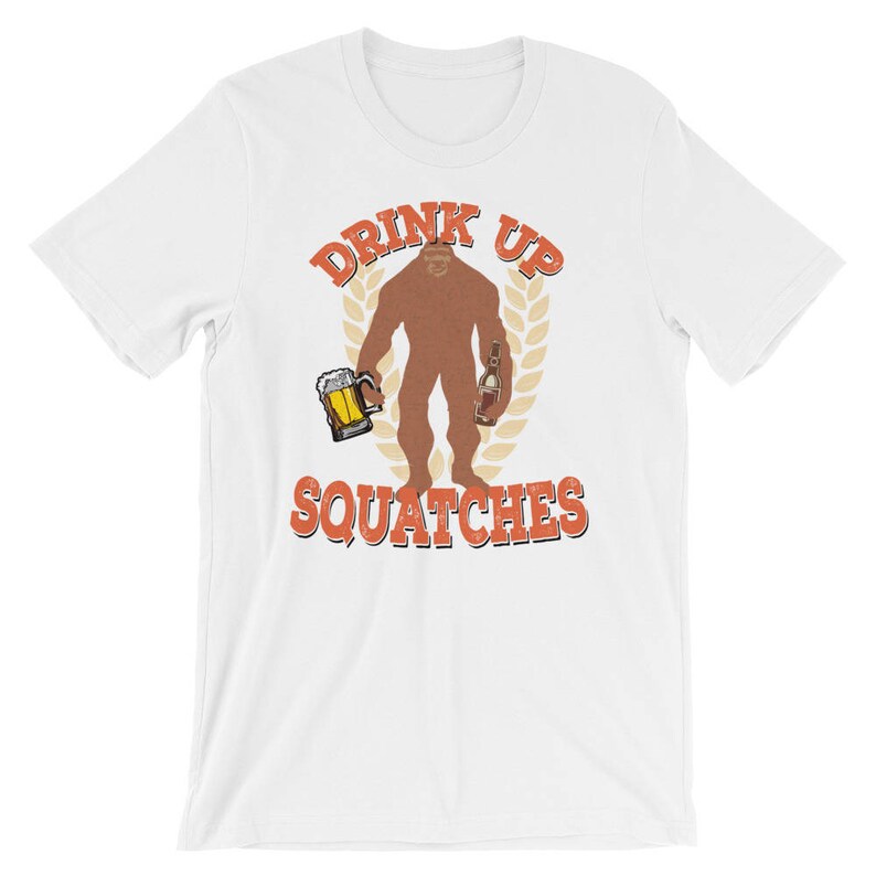 May include: White t-shirt with a brown Bigfoot holding a beer and a bottle. The text "Drink Up Squatchies" is printed in orange on the shirt.