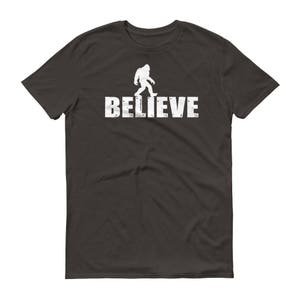 May include: Dark gray t-shirt with a white silhouette of Bigfoot walking and the text "BELIEVE" underneath.