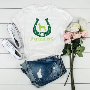 Puede incluir: Camiseta blanca con una herradura verde y un trébol verde. La herradura tiene una silueta verde de un perro en su interior. El texto "you lucky dog" está impreso debajo de la herradura.