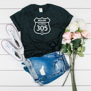 Puede incluir: Camiseta negra con un gráfico blanco de una señal de carretera vintage que dice "Miami, Florida 305 USA".