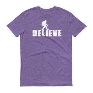 May include: Purple t-shirt with a white silhouette of Bigfoot walking and the word "BELIEVE" printed below.