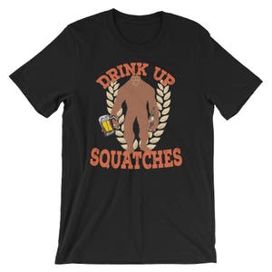 May include: Black t-shirt with a graphic of a Bigfoot holding a beer and a bottle. The text "Drink Up Squatchies" is printed in orange on the shirt.