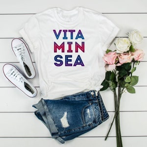 May include: White t-shirt with the text "VITA MIN SEA" in a gradient of pink, purple, and blue.