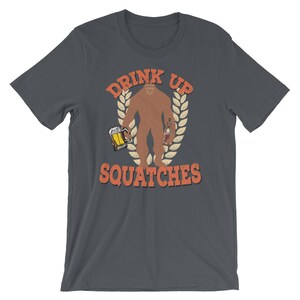 May include: Dark gray t-shirt with a graphic of a Bigfoot holding a beer and a bottle. The text "Drink Up Squatchees" is printed in orange on the shirt.