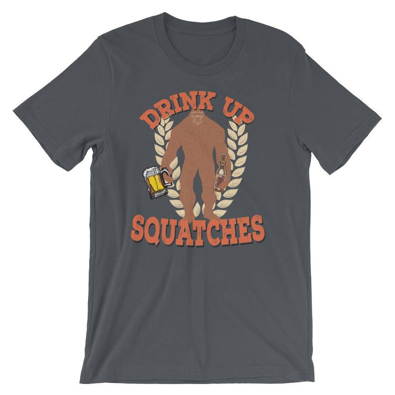 May include: Dark gray t-shirt with a graphic of a Bigfoot holding a beer and a bottle. The text "Drink Up Squatchees" is printed in orange on the shirt.