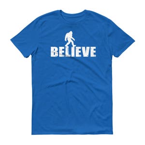 May include: A royal blue t-shirt with a white silhouette of Bigfoot walking and the word "BELIEVE" printed below.