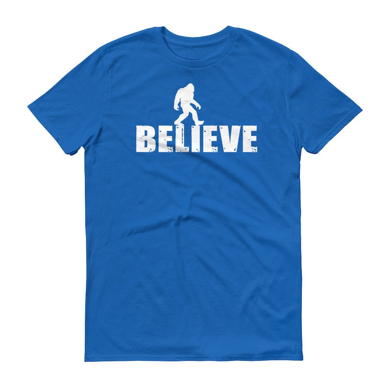 May include: A royal blue t-shirt with a white silhouette of Bigfoot walking and the word "BELIEVE" printed below.