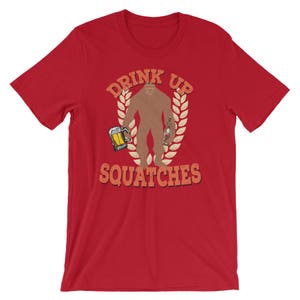 May include: Red t-shirt with a graphic of a Bigfoot holding a beer and a bottle. The text "Drink Up Squatche" is printed on the shirt.
