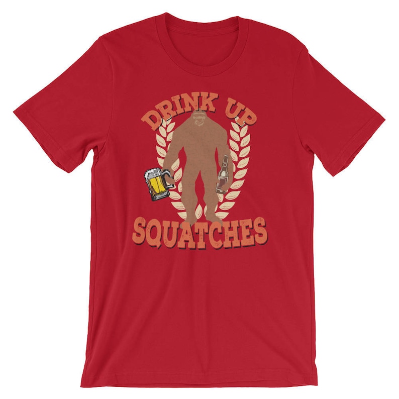 May include: Red t-shirt with a graphic of a Bigfoot holding a beer and a bottle. The text "Drink Up Squatche" is printed on the shirt.