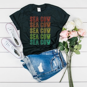 Puede incluir: Una camiseta negra con el texto "SEA COW" repetido cinco veces en un degradado arcoíris. El texto está en una fuente de estilo retro. La camiseta está colocada sobre una superficie de madera blanca con un par de zapatillas blancas y un ramo de rosas rosas.