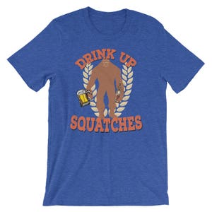 May include: A blue t-shirt with the text "Drink Up Squatche" in orange. The graphic features a brown Bigfoot holding a beer and a bottle.