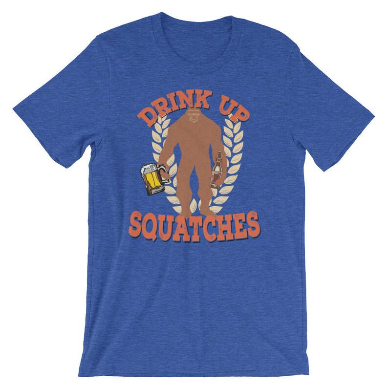 May include: A blue t-shirt with the text "Drink Up Squatche" in orange. The graphic features a brown Bigfoot holding a beer and a bottle.