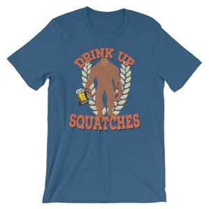 May include: A blue t-shirt with a graphic of a sasquatch holding a beer and a bottle. The text "Drink Up Squatche" is printed on the shirt.