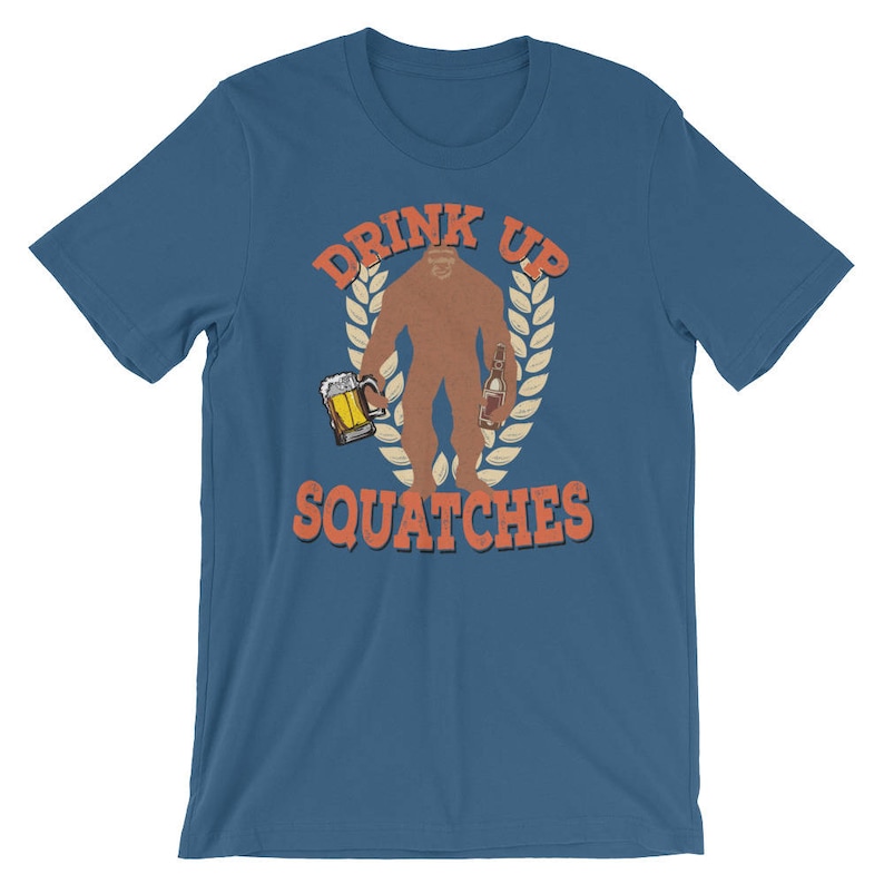 May include: A blue t-shirt with a graphic of a sasquatch holding a beer and a bottle. The text "Drink Up Squatche" is printed on the shirt.