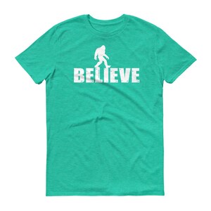 May include: A teal blue t-shirt with a white silhouette of Bigfoot walking and the word "BELIEVE" printed below it.