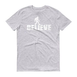 May include: Gray short-sleeved t-shirt with a white silhouette of Bigfoot walking and the word "BELIEVE" printed below.
