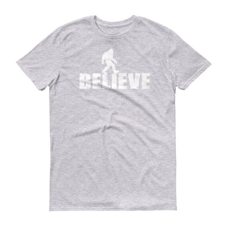 May include: Gray short-sleeved t-shirt with a white silhouette of Bigfoot walking and the word "BELIEVE" printed below.