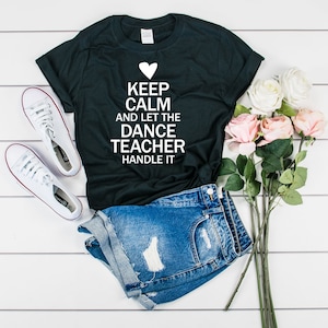 Könnte beinhalten: Schwarzes T-Shirt mit weißem Text, der "Keep Calm and Let the Dance Teacher Handle It" lautet. Über dem Text befindet sich ein Herzsymbol. Das Shirt liegt auf einer weißen Oberfläche mit einem Paar weißen Sneakers und einer Jeans-Shorts.