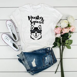 May include: White t-shirt with a black husky dog wearing sunglasses and the text "Husky Squad" printed on it. The shirt is laid out on a white wooden surface with a pair of white sneakers and a pair of blue denim shorts.