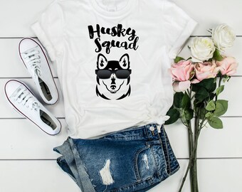 May include: White t-shirt with a black husky dog wearing sunglasses and the text "Husky Squad" printed on it. The shirt is laid out on a white wooden surface with a pair of white sneakers and a pair of blue denim shorts.