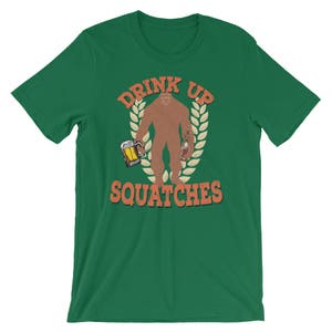 May include: Green t-shirt with a cartoon Bigfoot holding a beer and a bottle. The text "Drink Up Squatchies" is printed on the shirt.