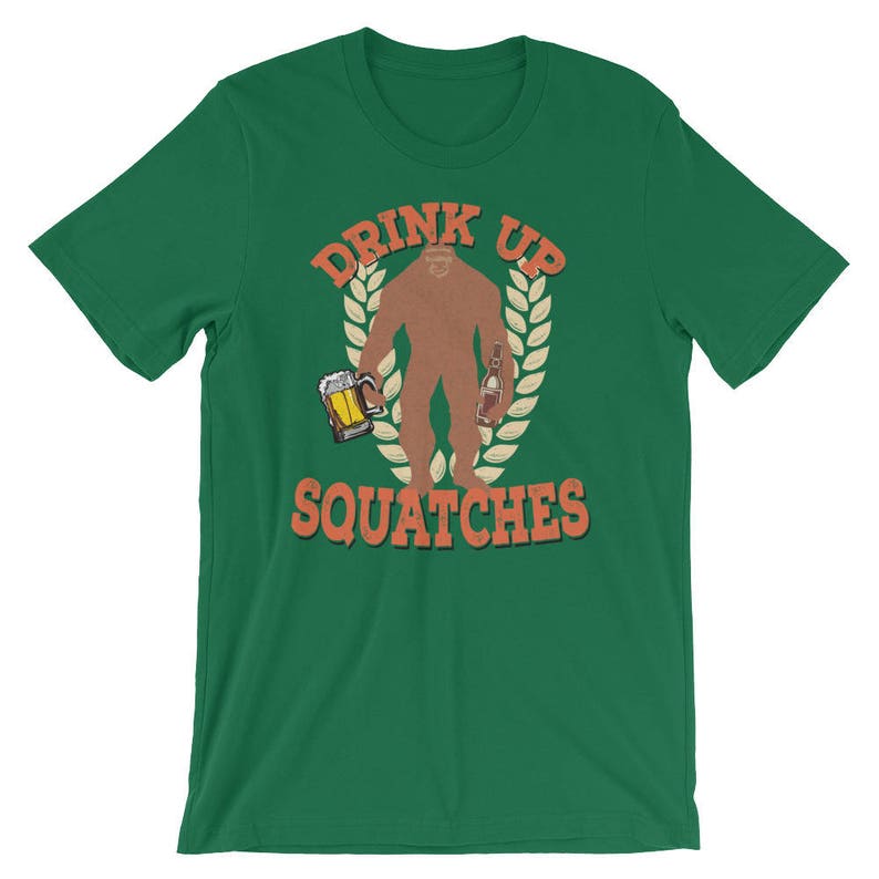May include: Green t-shirt with a cartoon Bigfoot holding a beer and a bottle. The text "Drink Up Squatchies" is printed on the shirt.