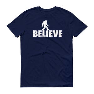 May include: A navy blue t-shirt with a white silhouette of Bigfoot walking and the word "BELIEVE" printed below it.