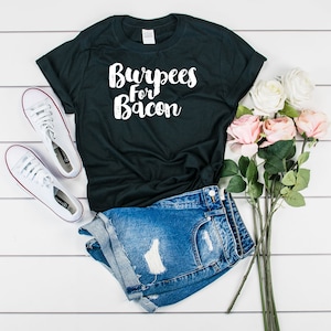 May include: A black t-shirt with the text "Burpees For Bacon" in white lettering. The shirt is paired with denim shorts and white sneakers. A bouquet of pink and white roses is in the background.