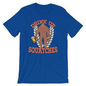 May include: A royal blue t-shirt with a graphic of a Bigfoot holding a beer and a bottle of liquor. The graphic is surrounded by a wreath of leaves and the text "Drink Up Squatchies".