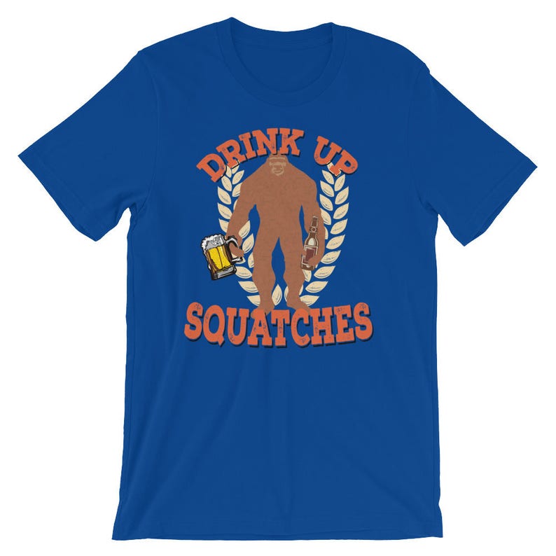 May include: A royal blue t-shirt with a graphic of a Bigfoot holding a beer and a bottle of liquor. The graphic is surrounded by a wreath of leaves and the text "Drink Up Squatchies".