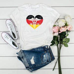 May include: White t-shirt with a heart-shaped design featuring the German flag colors and a medical symbol. The letters "R" and "T" are visible within the heart. The shirt is paired with denim shorts and white sneakers, with a bouquet of pink and white flowers.