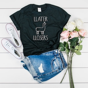 Könnte beinhalten: Schwarzes T-Shirt mit einer weißen Lama-Illustration und dem Aufdruck "LLATER LLOSERS" auf der Vorderseite. Das Shirt wird mit Jeans-Shorts und weißen Sneakern kombiniert.