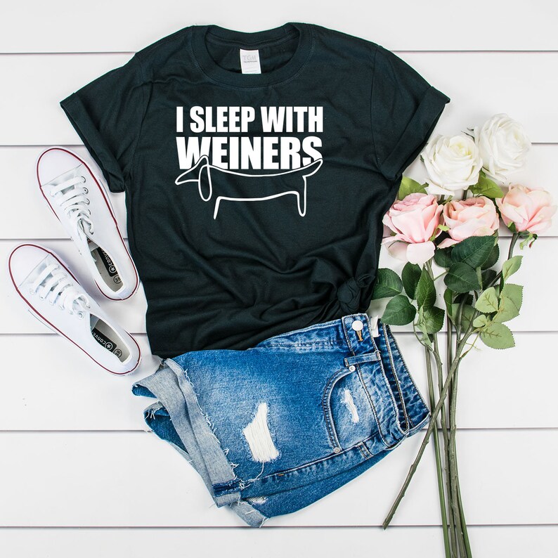 Pode incluir: Uma camiseta preta com texto branco que diz "I sleep with weiners" e um desenho simples de um dachshund. A camiseta &eacute; combinada com shorts jeans e t&ecirc;nis brancos.