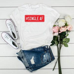 May include: White t-shirt with a red rectangular graphic that says "#SINGLE AF" on it. The shirt is paired with denim shorts and white sneakers.