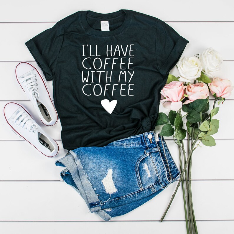 May include: Black t-shirt with white text that reads "I'll have coffee with my coffee" and a white heart. The shirt is paired with denim shorts and white sneakers.