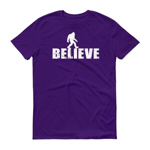 May include: Purple t-shirt with a white silhouette of Bigfoot walking and the word "BELIEVE" printed below.