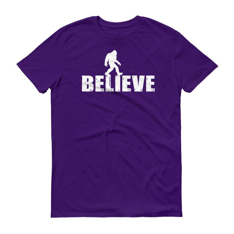 May include: Purple t-shirt with a white silhouette of Bigfoot walking and the word "BELIEVE" printed below.