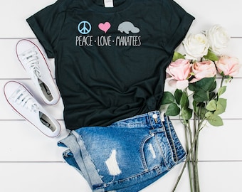 May include: Black t-shirt featuring a peace sign, heart, and manatee graphic. The text "PEACE - LOVE - MANATEES" is printed below the graphic. The shirt is styled with denim shorts and white trainers.