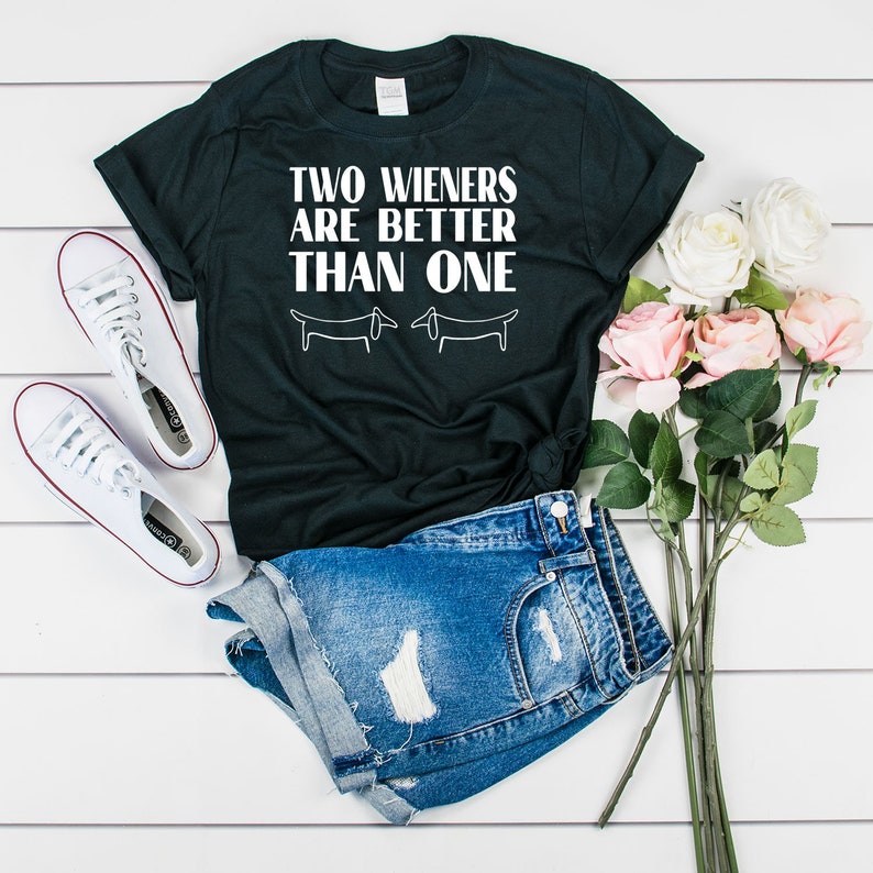 Puede incluir: Camiseta negra con texto blanco que dice "TWO WIENERS ARE BETTER THAN ONE" y dos ilustraciones de perros dachshund. La camiseta est&aacute; sobre una superficie de madera blanca con un par de zapatillas blancas, pantalones cortos de mezclilla y flores rosas.