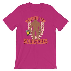 May include: A pink t-shirt with a graphic of a Bigfoot holding a beer and a bottle. The text "Drink Up Squatchies" is printed in orange and yellow.
