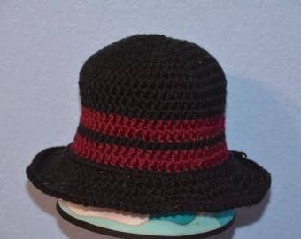 May include: A black crochet hat with a wide brim and a burgundy band. The hat is handmade and appears to be made of yarn. The hat is sitting on a light blue surface.