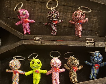 May include: A collection of eight colorful voodoo doll keychains. The dolls are made of yarn and have different colored eyes and mouths. They are all wearing red hearts. The keychains are displayed in a wooden box.