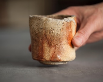 May include: A handcrafted ceramic cup with a textured surface and an irregular shape. The cup displays a mix of beige, brown, and orange tones, with a slightly rough texture. The cup is held by a hand, highlighting its form and size.