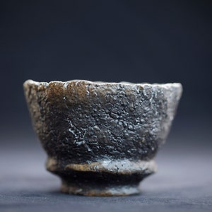 K&ouml;nnte beinhalten: Eine kleine, schwarze Keramiksch&uuml;ssel mit einer rauen, strukturierten Oberfl&auml;che. Die Sch&uuml;ssel hat eine breite, abgerundete &Ouml;ffnung und einen leicht ausgestellten Boden.