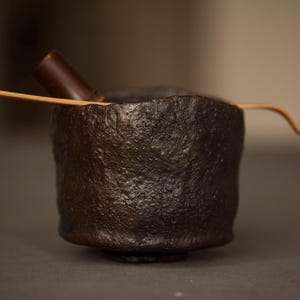 May include: A dark brown, textured ceramic tea bowl with a bamboo whisk and scoop. The bowl has a rough, organic texture and a small, rounded base. The whisk and scoop are light brown.