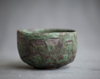 May include: A small, green ceramic bowl with a textured, mottled surface. The bowl has a slightly rounded shape and a wide rim.