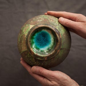 Op de afbeelding: Een groene en bruine keramische kom met een turquoise blauwe glazuur aan de binnenkant. De kom wordt vastgehouden door twee handen.