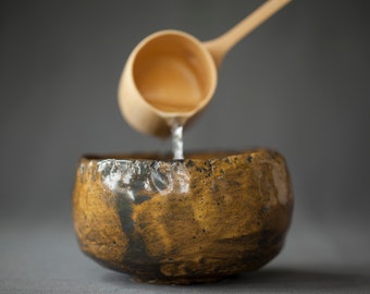 May include: A wooden ladle pours water into a brown ceramic bowl. The bowl has a textured, uneven surface.