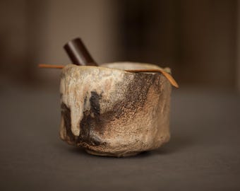 Puede incluir: Un cuenco de té de cerámica texturizada con una forma orgánica irregular, una base de color marrón oscuro y un esmalte de color crema que gotea por los lados. Una fina cuchara de té de madera descansa sobre el borde y un objeto cilíndrico marrón oscuro está dentro.